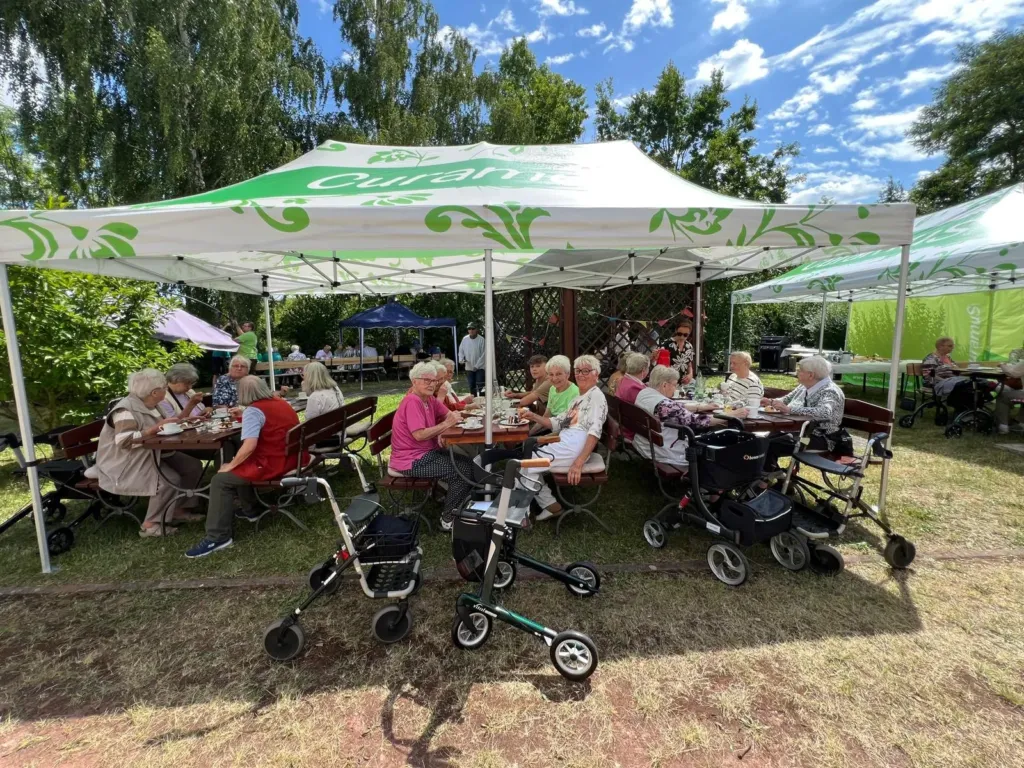 Seniorinnen und Senioren sitzen unter Curamus-Pavillons im Garten beim Sommerfest, Rollatoren stehen bereit